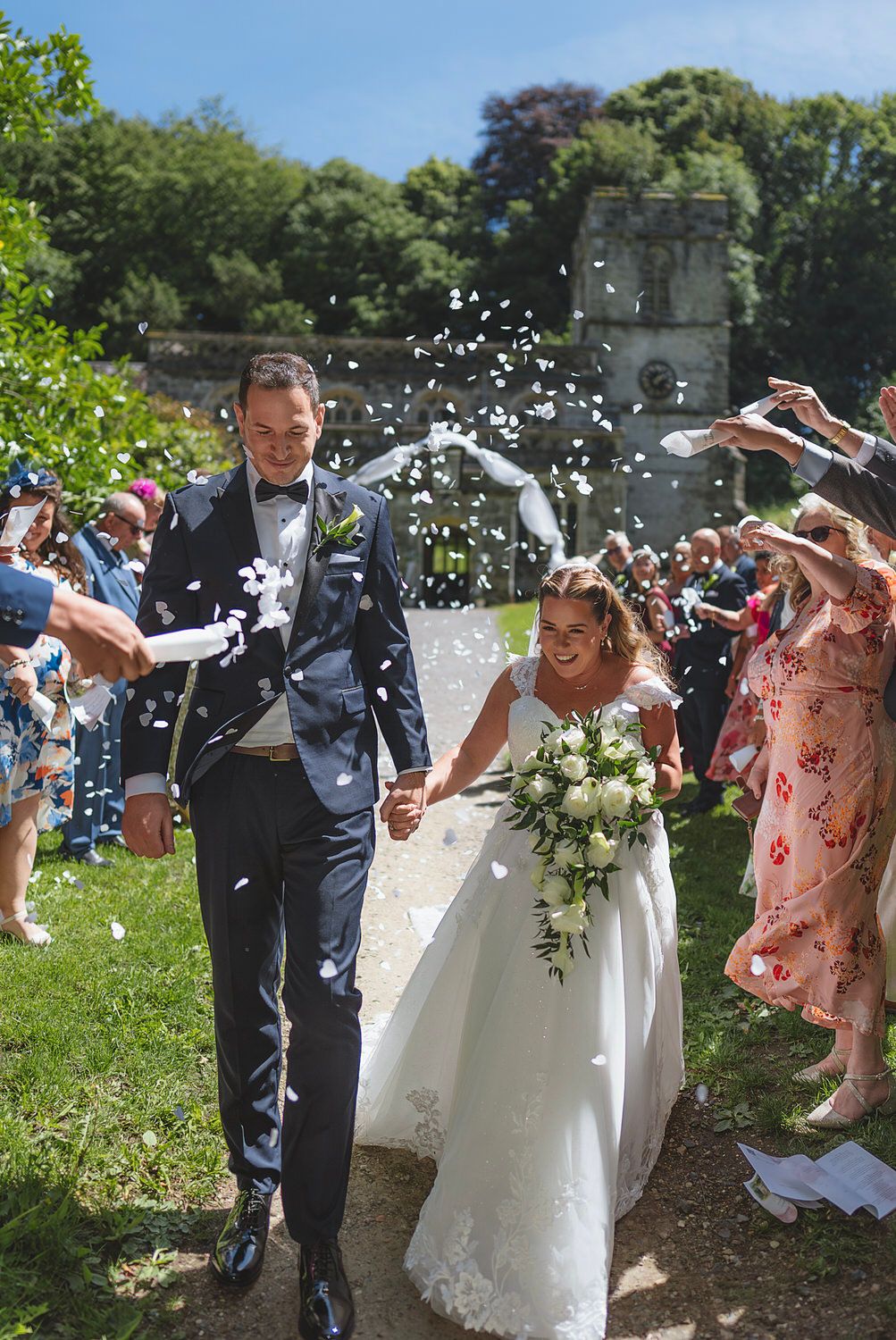 Bride and groom walk through confetti shower, guests celebrating outdoor wedding ceremony on sunny day.