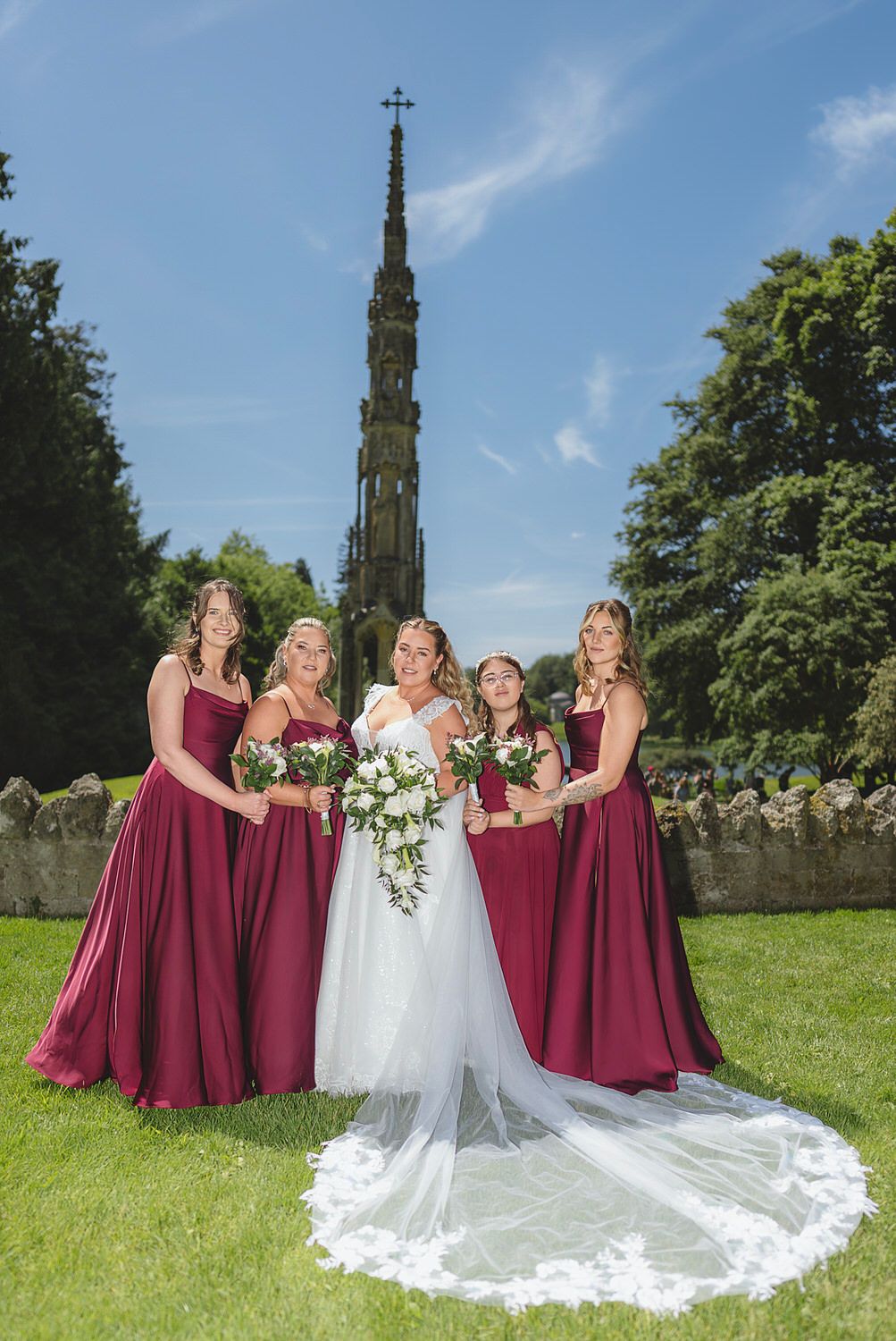 Bride in white gown with four bridesmaids in burgundy dresses holding bouquets outdoors near a stone church spire.