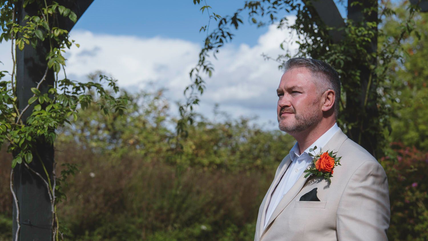 Groom in beige suit with orange boutonniere standing under floral arch at outdoor wedding ceremony.