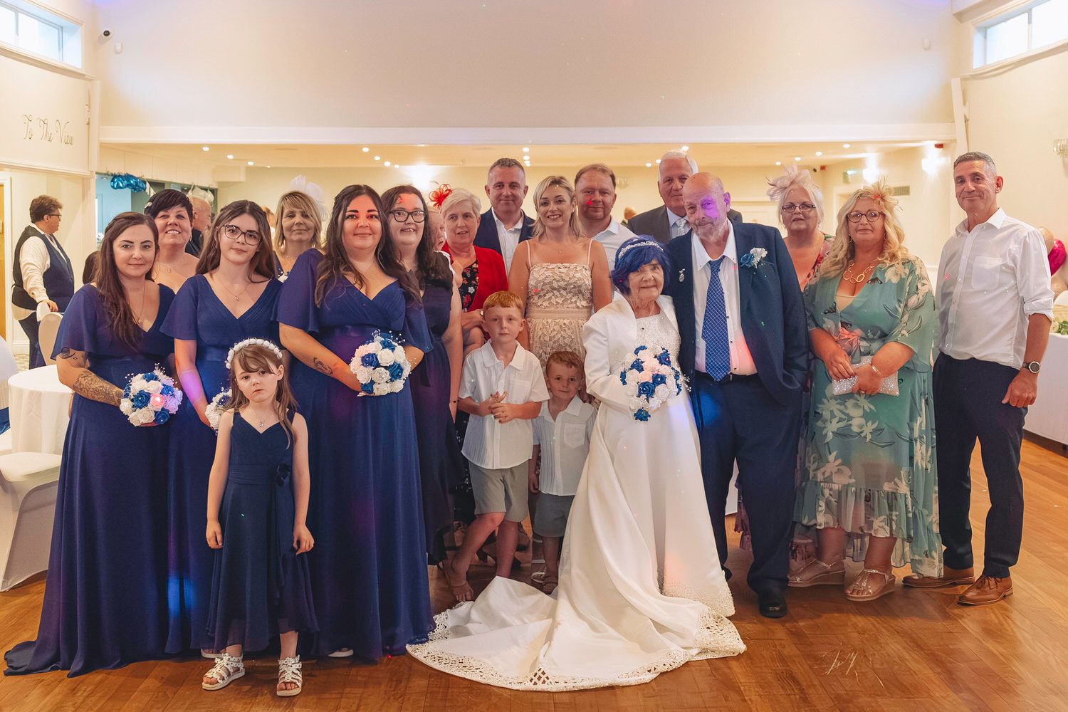 Wedding party poses together in a hall, bride in white gown with blue bouquet, bridesmaids in navy dresses.