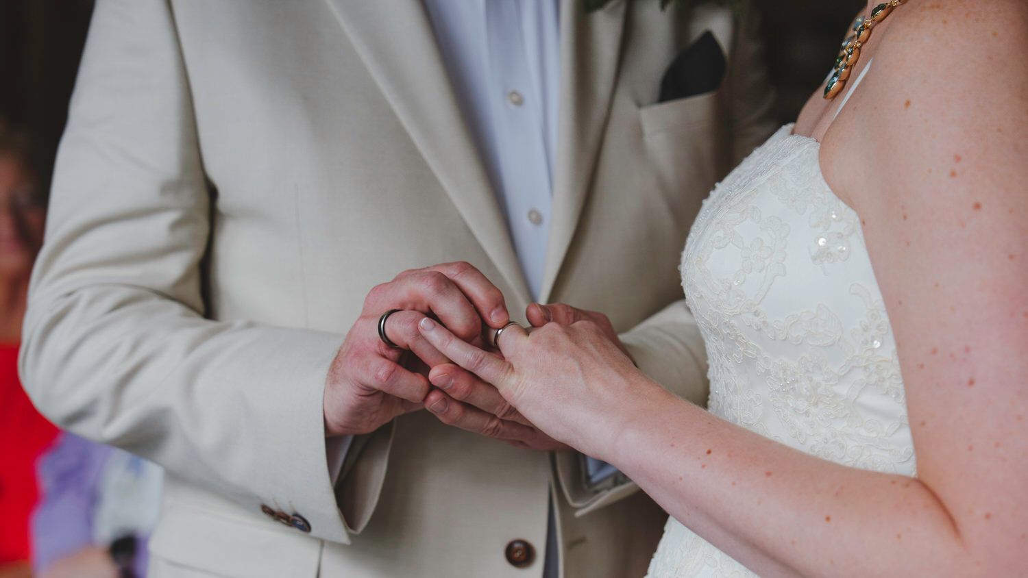 Bride placing a wedding ring on groom's finger during ceremony, both dressed in white and beige attire.