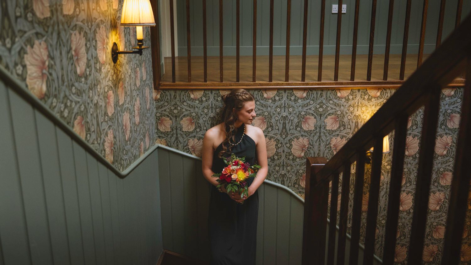 Bridesmaid in dark gown holding colorful bouquet on staircase with floral wallpaper and warm lamp light.