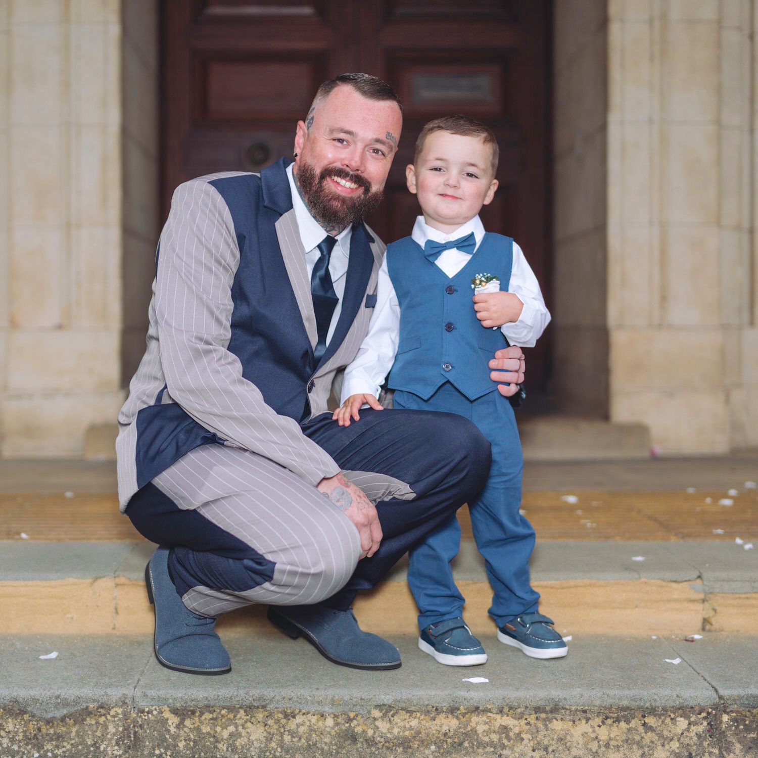 A man in a gray suit crouches beside a young boy in a blue vest and bow tie on stone steps.