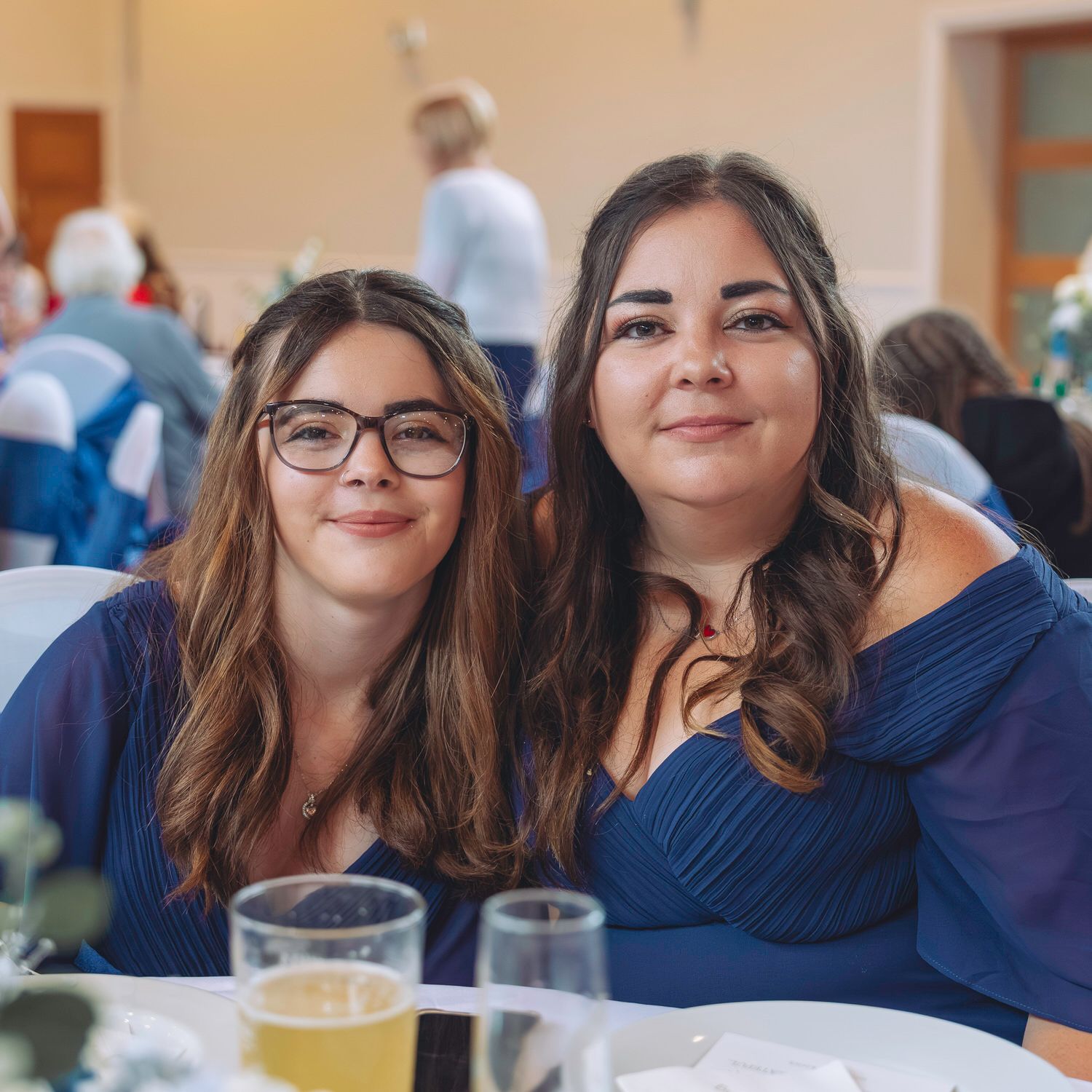 Two women in navy blue dresses smiling at a formal event, seated at a decorated table.