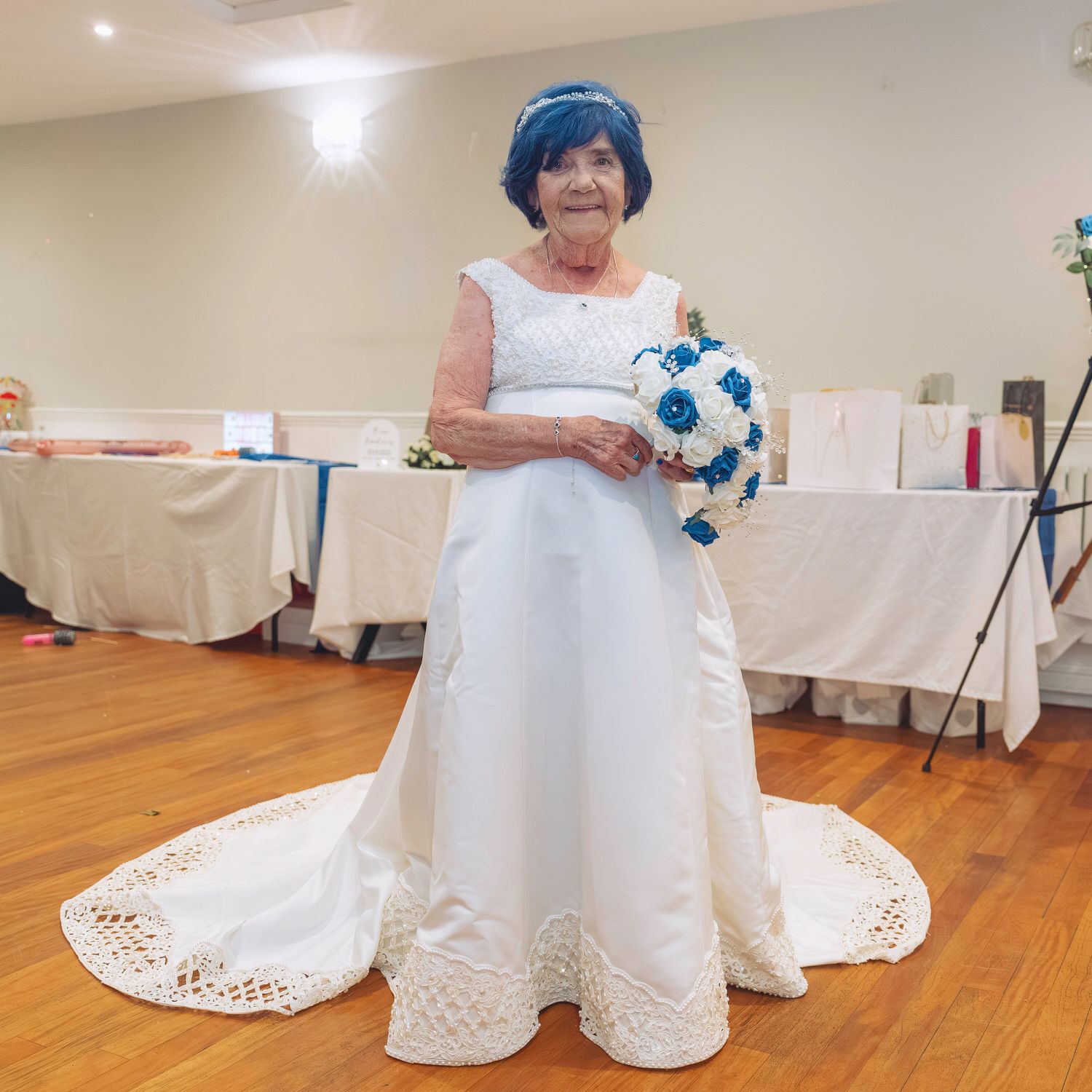 Elderly woman in white wedding gown with lace trim holding blue and white bouquet, wearing blue headpiece.