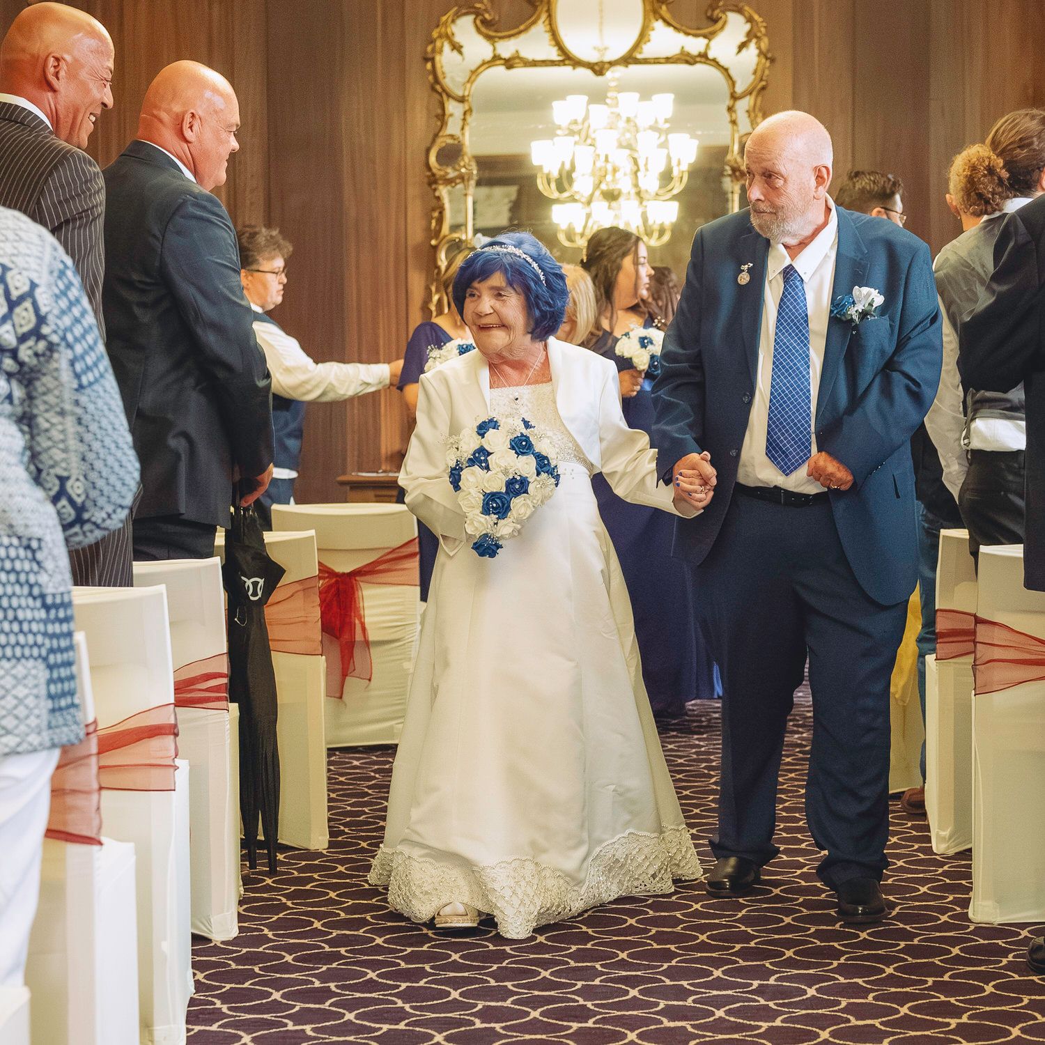 Elderly couple walking down wedding aisle, bride in white gown with blue hat holding bouquet, groom in navy suit.