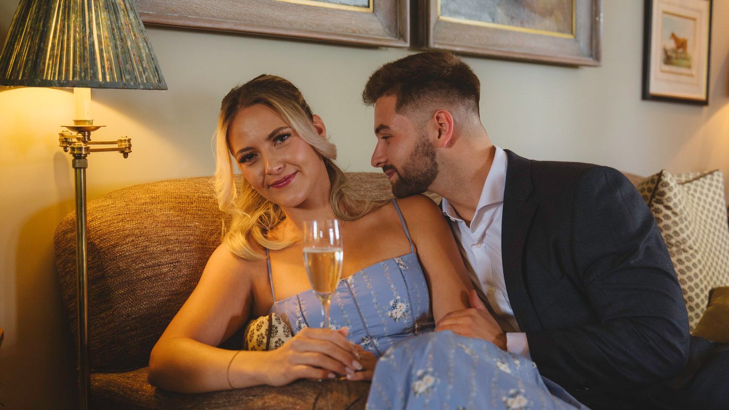 Romantic couple sharing champagne on a cozy sofa, woman in blue dress smiling at man in suit.