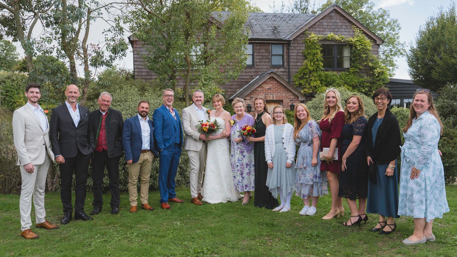 Wedding party posing outdoors in front of a brick house, bride in white gown holding bouquet surrounded by guests.