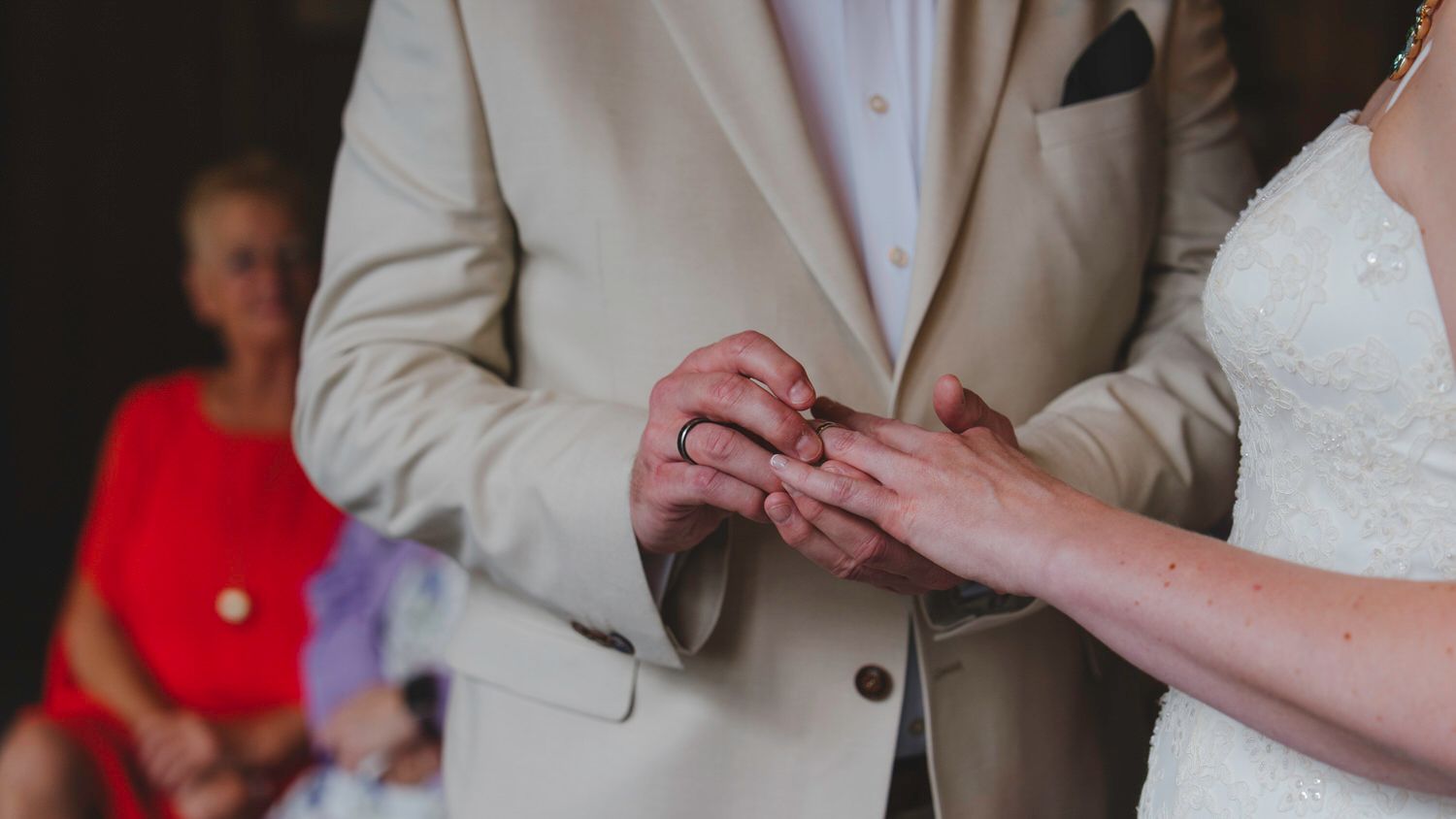 Groom placing wedding ring on bride's finger during ceremony, with guest in red dress visible in background.