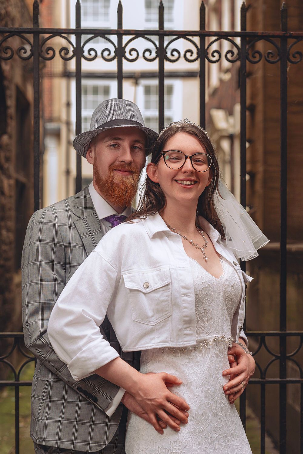 A smiling bride and groom pose before iron gates, she in white dress and tiara, he in grey plaid suit and fedora hat.