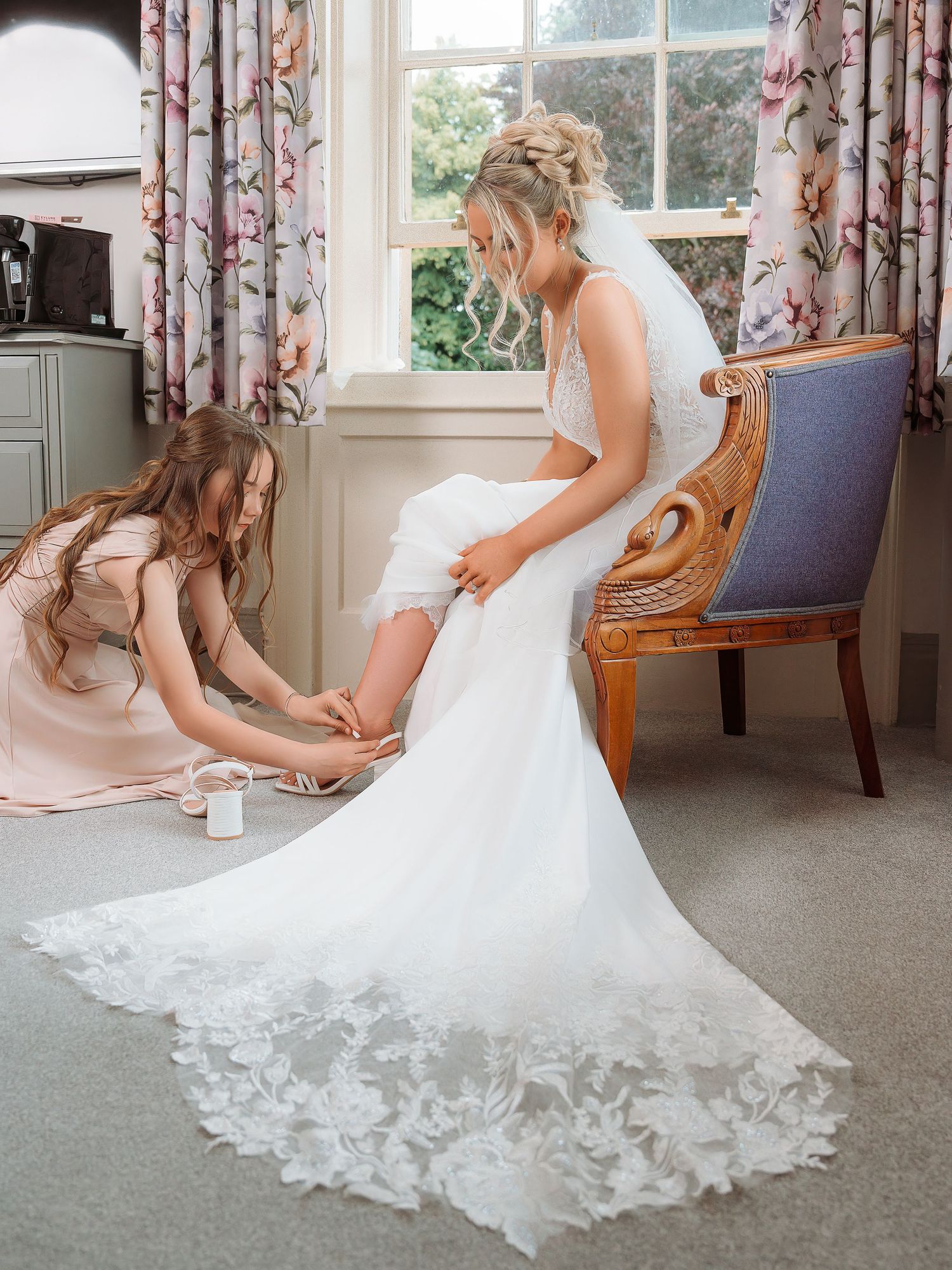 Bride in lace wedding gown sits as bridesmaid in blush dress helps with shoes near a window with floral curtains.
