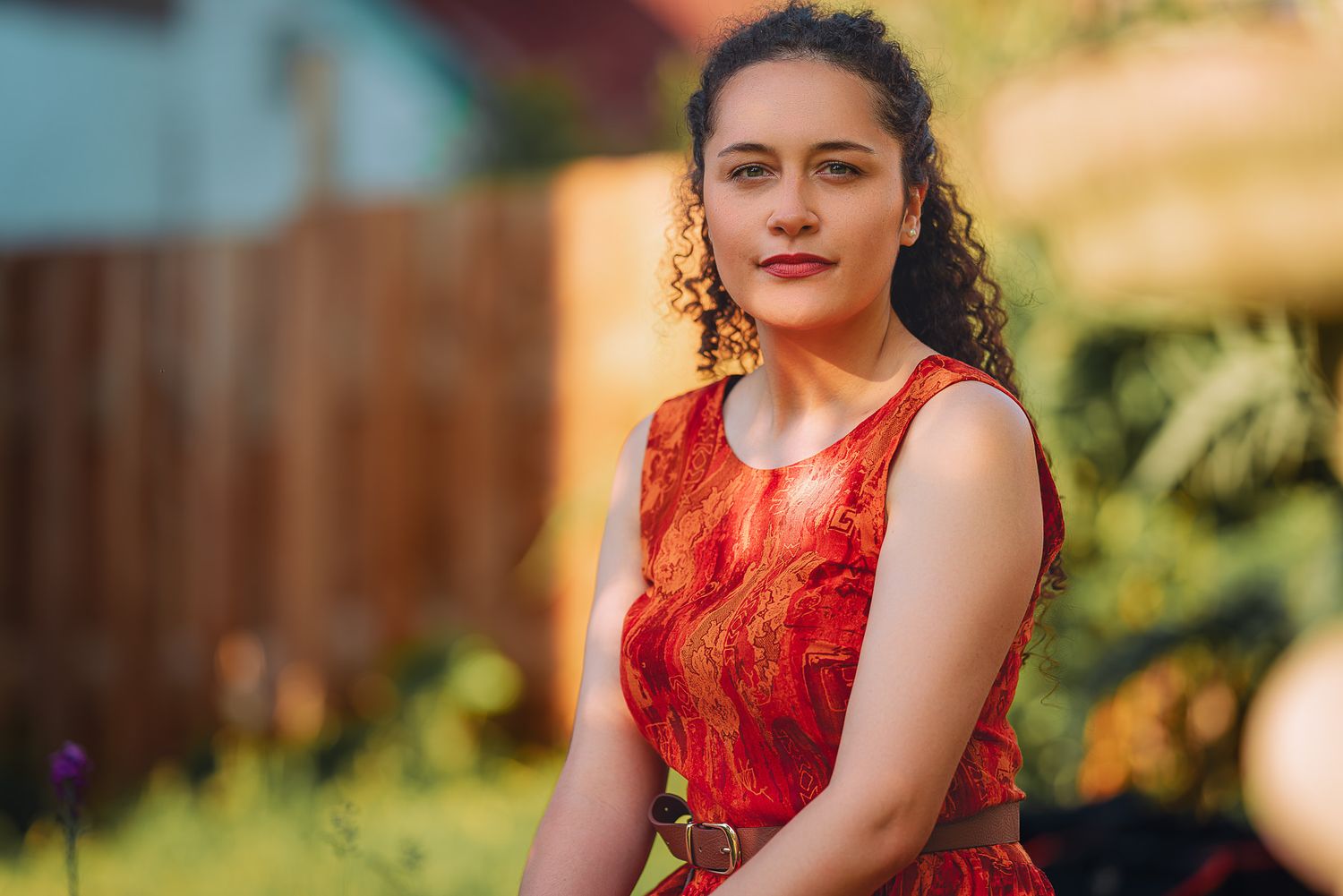 Young woman in red lace dress poses in sunlit garden with warm bokeh background.