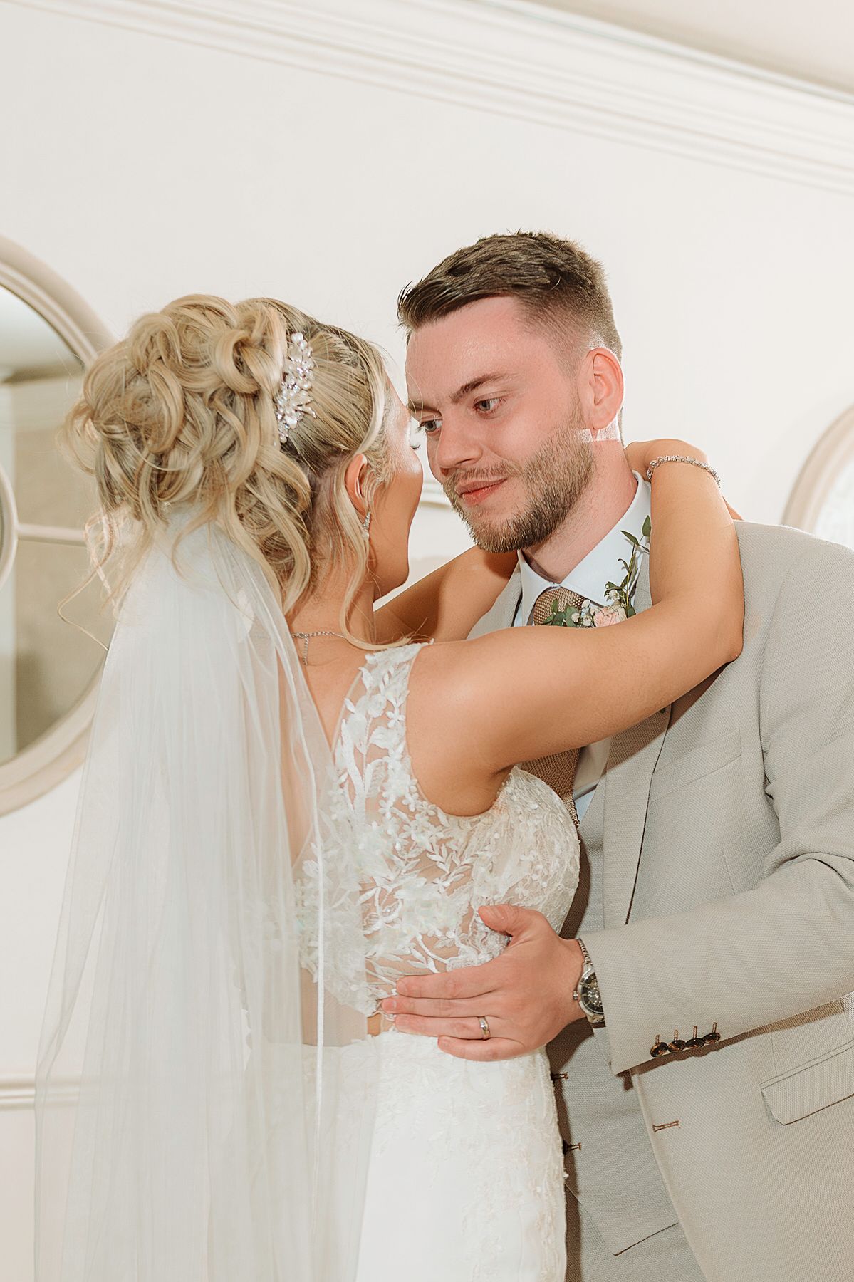 Groom in grey suit embracing bride in lace wedding dress indoors, sharing a tender moment.