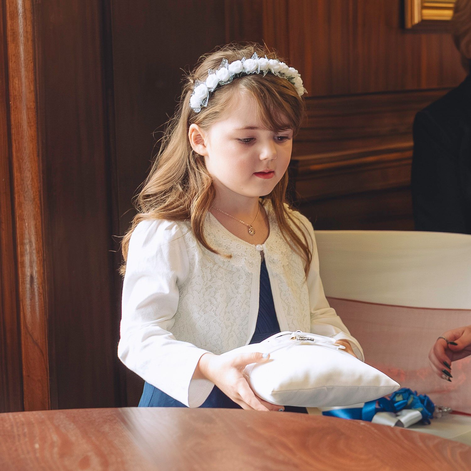 Young girl in white jacket and floral headband holds a ring pillow at a wedding ceremony.