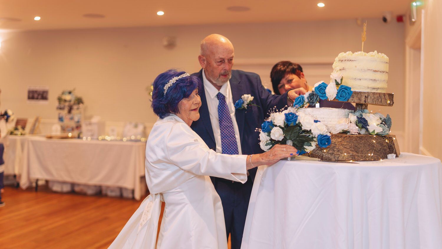 Elderly couple cutting wedding cake at reception, decorated with blue flowers, in elegant white banquet hall.