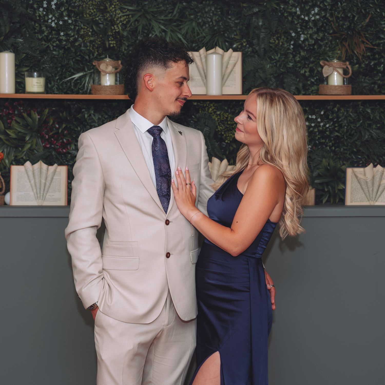 Couple in elegant attire, man in beige suit and woman in navy dress, sharing a romantic moment indoors.