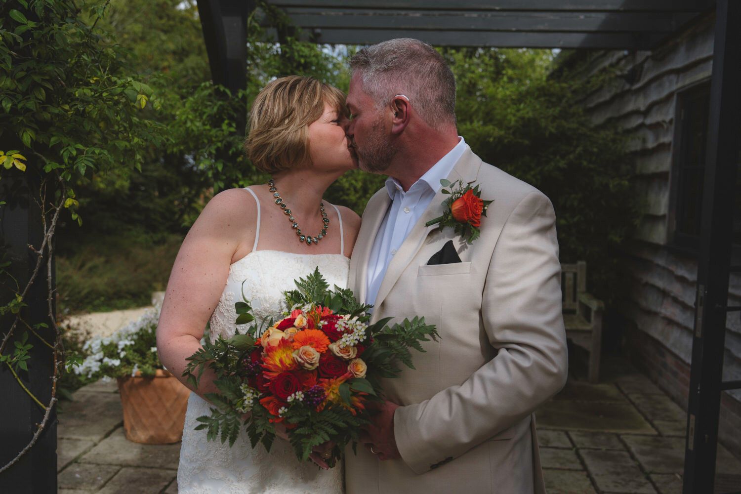 Bride and groom share a kiss outdoors, bride holds vibrant orange and red bouquet, groom wears beige suit with boutonniere.