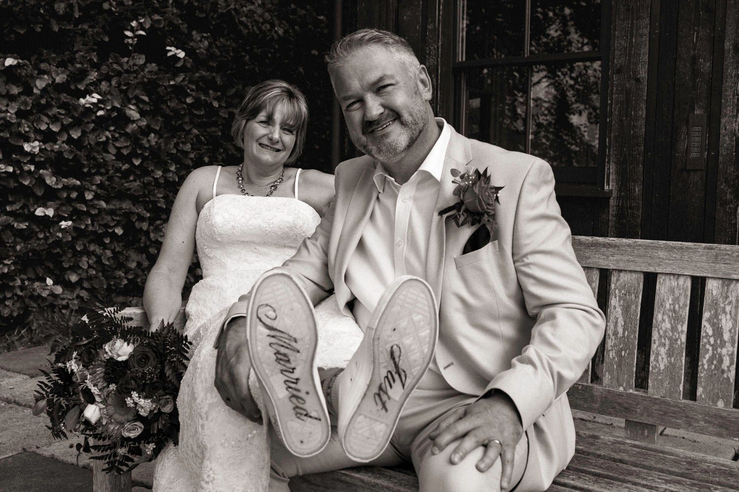 Bride and groom laugh on a bench showing customized wedding shoe soles in black and white photo.