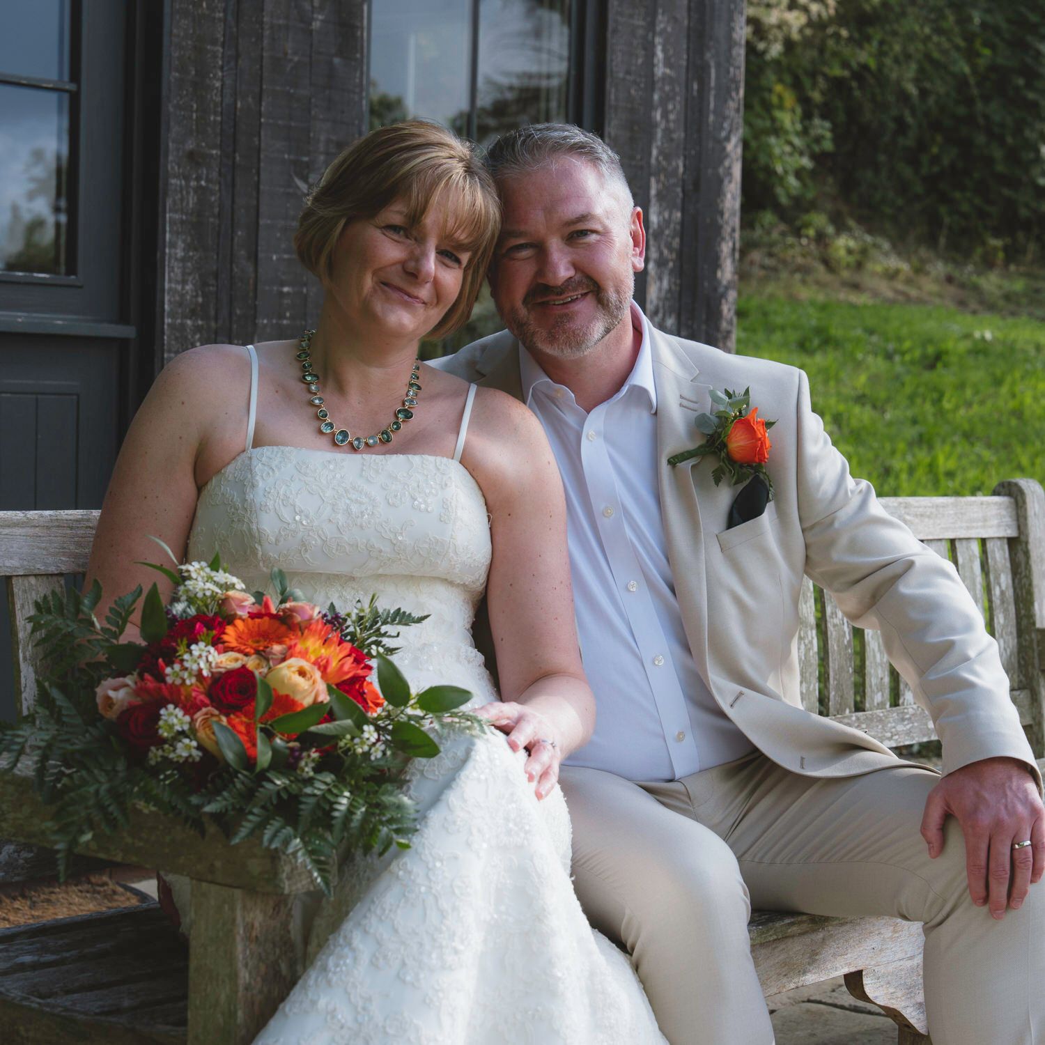Happy bride and groom sitting on a wooden bench, bride holding colorful bouquet, groom wearing boutonniere, outdoor wedding.