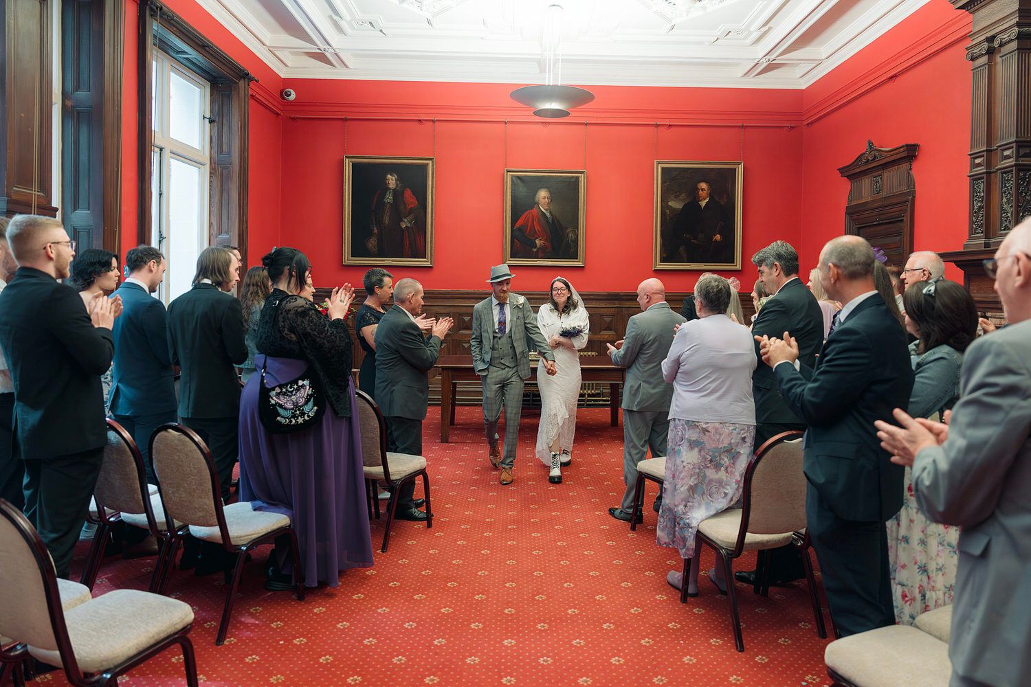 Newlywed couple walks through applauding guests in an elegant red-walled room during a wedding reception ceremony.