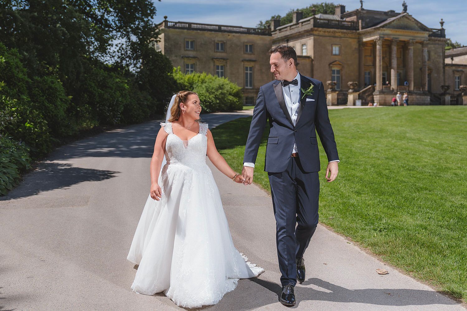 Bride and groom walk hand-in-hand on a sunlit path outside a grand estate on their wedding day.