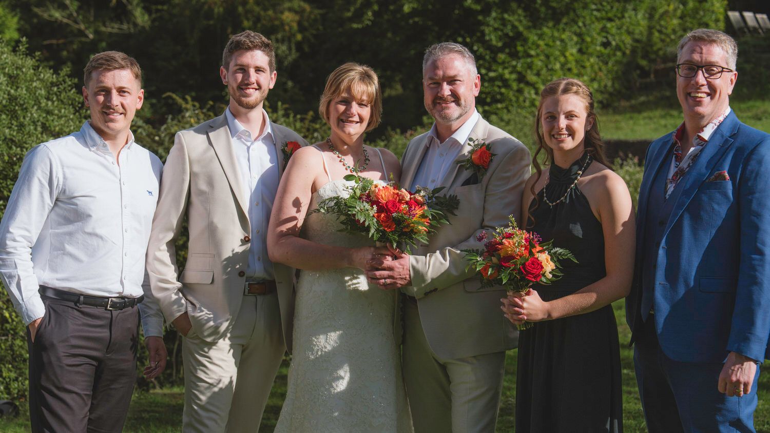 Wedding group photo with bride and groom holding bouquets, surrounded by four guests outdoors.