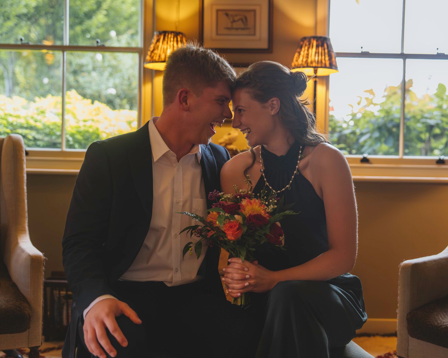 Couple sharing romantic moment with colorful bouquet indoors, leaning foreheads together near sunlit window.