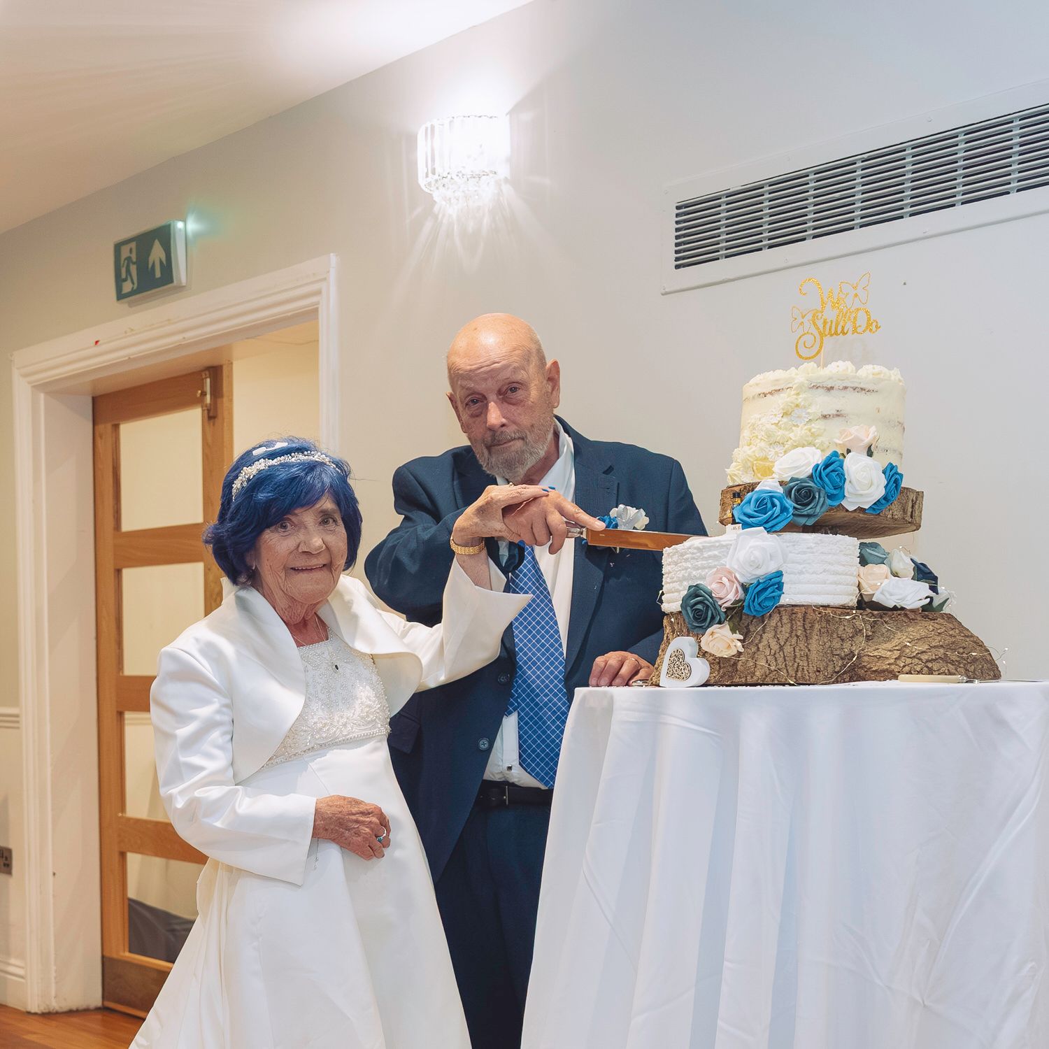 Elderly couple cutting their wedding cake together, bride in white dress and groom in suit with blue tie.