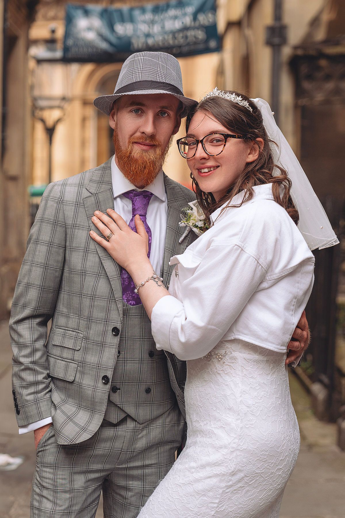 Groom in grey plaid suit and hat embraces bride in white lace dress with veil in vintage street.