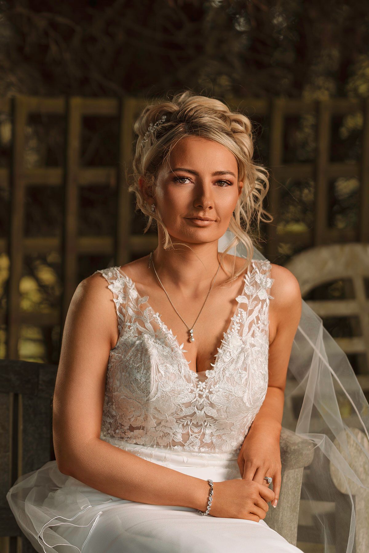 Elegant blonde bride in lace V-neck wedding dress with veil and jewelry seated outdoors.