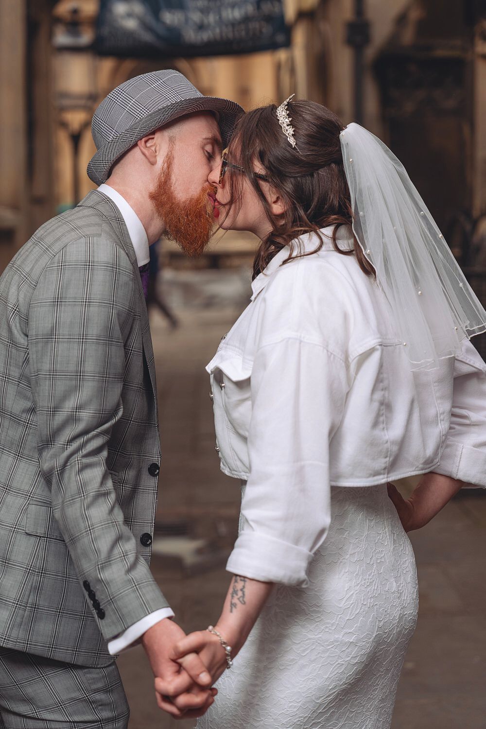 A bearded groom in a plaid suit and hat kisses his bride in a white dress and veil while holding hands outdoors.