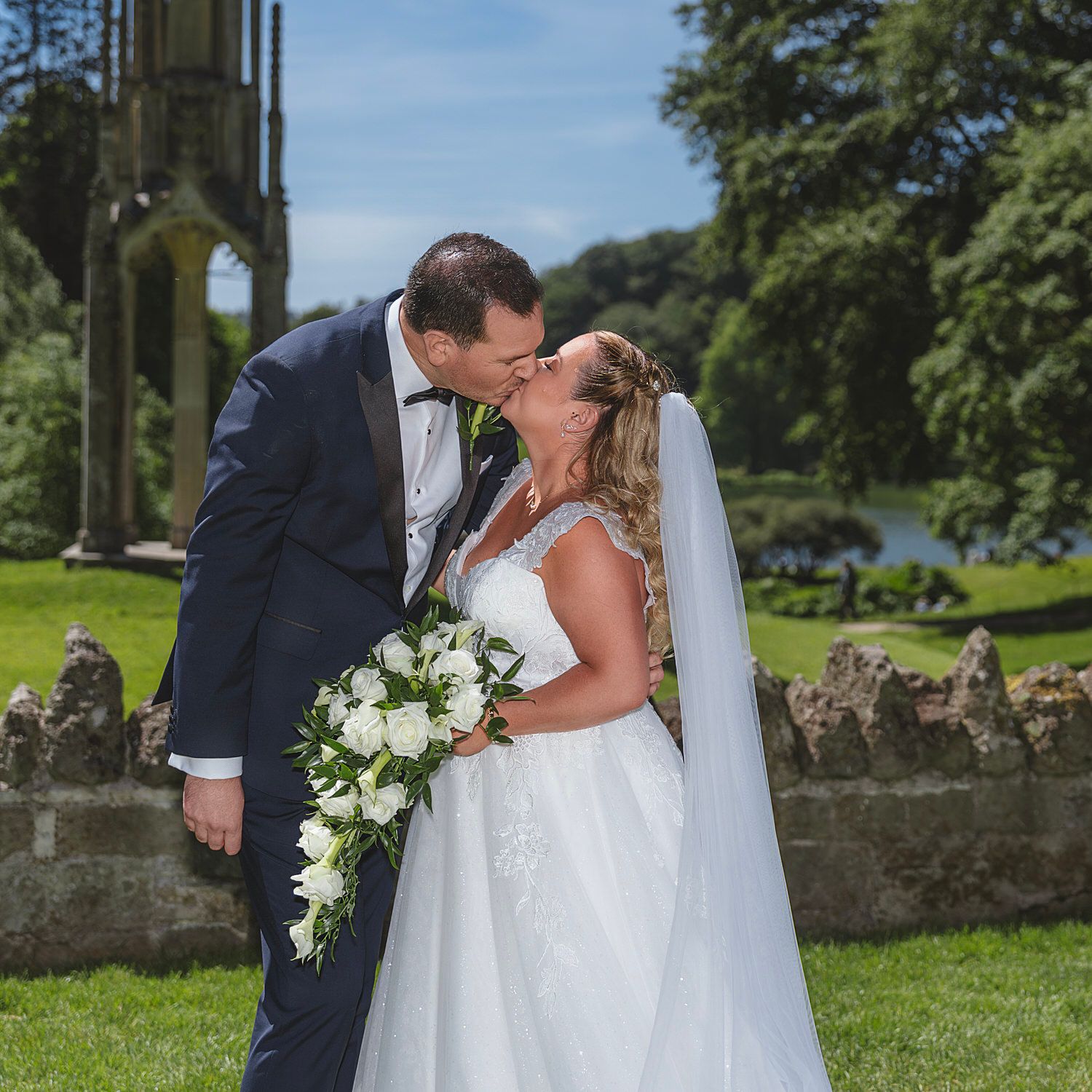 A bride and groom share a romantic kiss outdoors, with the bride holding a white bouquet, surrounded by lush greenery.
