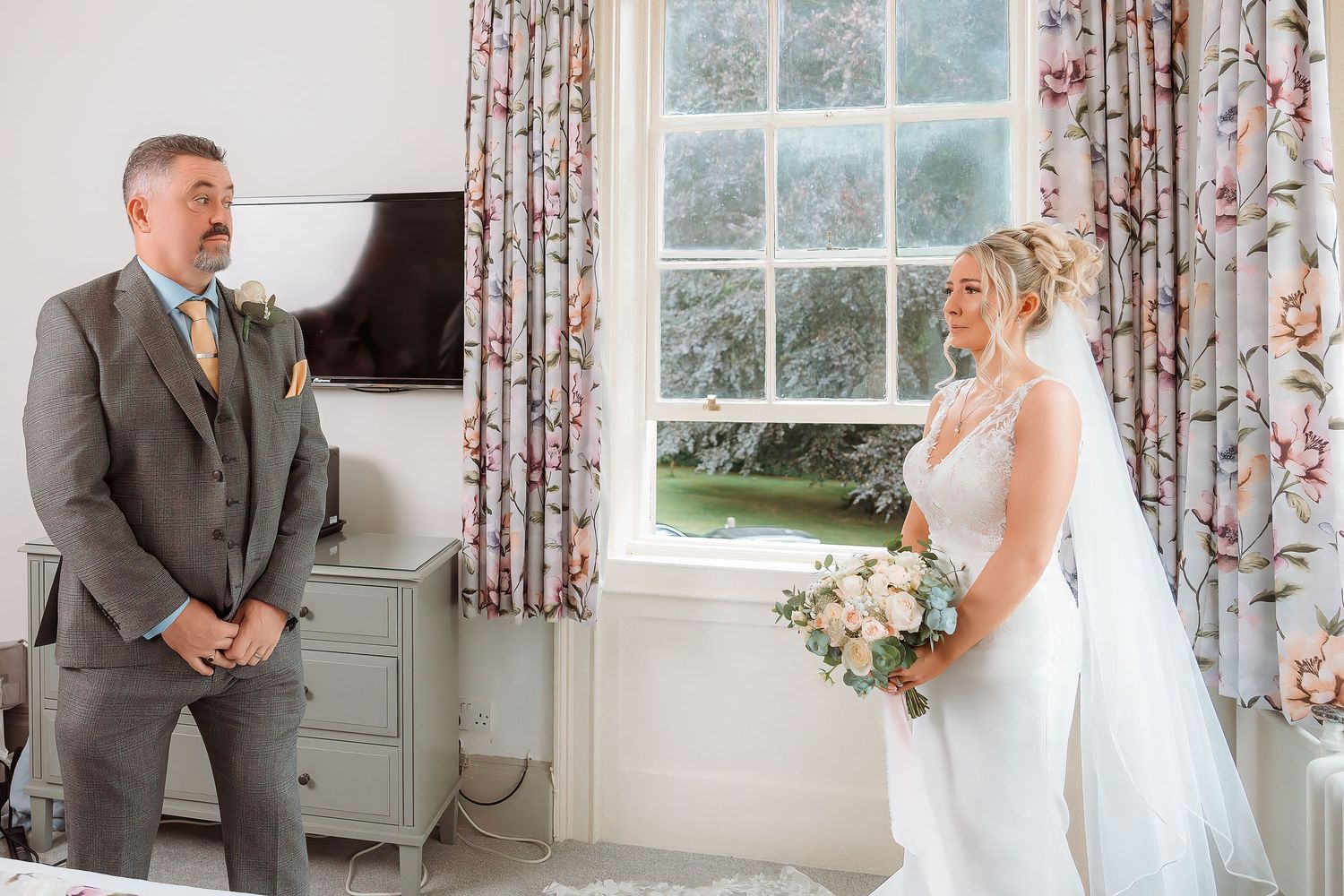Bride and groom share an emotional first look moment in a bright room with floral curtains, bride holding a white bouquet.