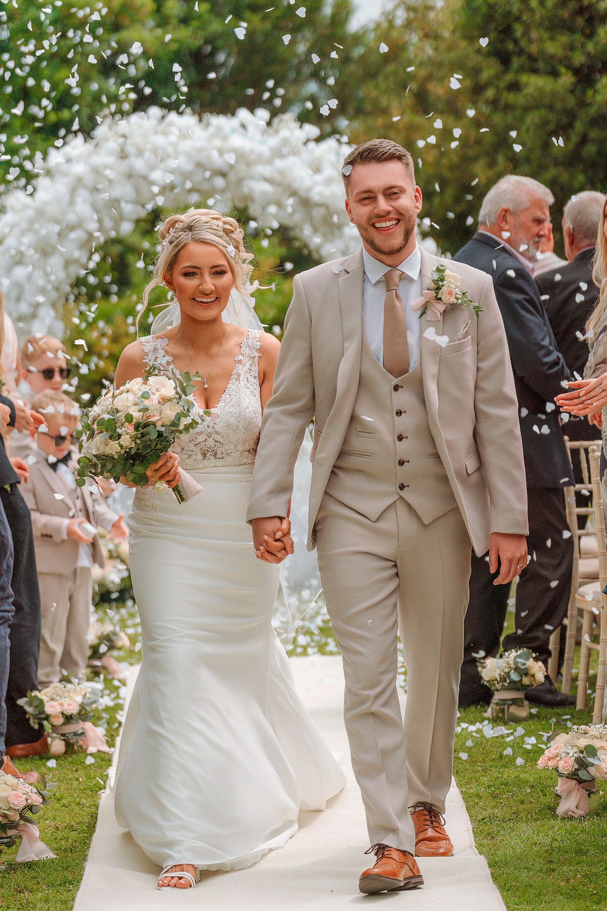 Newlyweds walking down aisle hand in hand as guests throw white confetti at outdoor wedding.