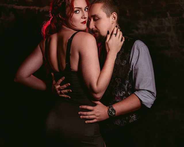 A couple in an intimate embrace, woman with red hair in a dark dress, man in a vest and watch, moody dark background.