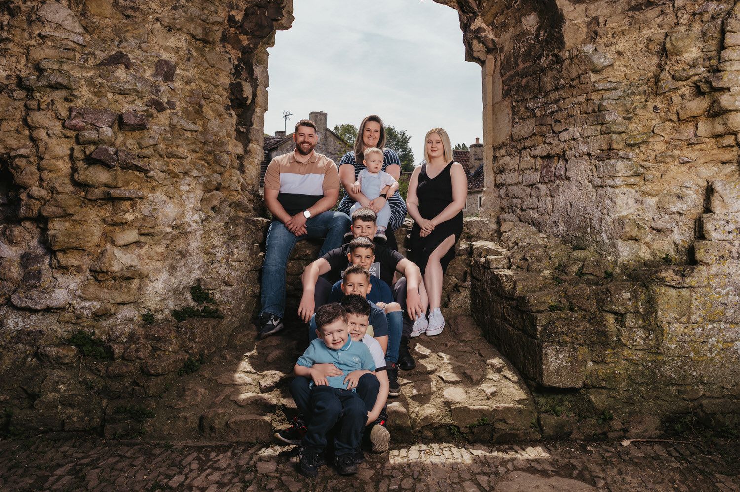 Family group portrait posed in ancient stone ruins archway, multiple generations smiling together outdoors.
