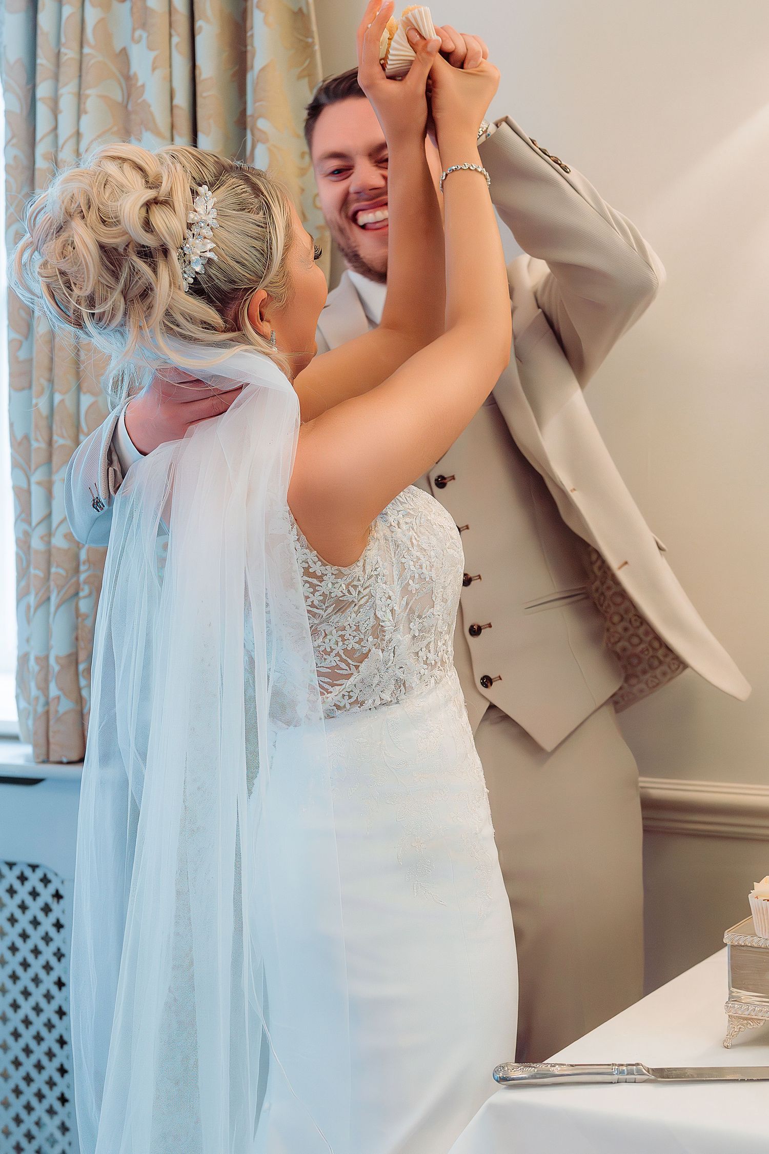 Bride and groom share a joyful first dance, with the bride in a lace gown and veil, groom in a tan suit.