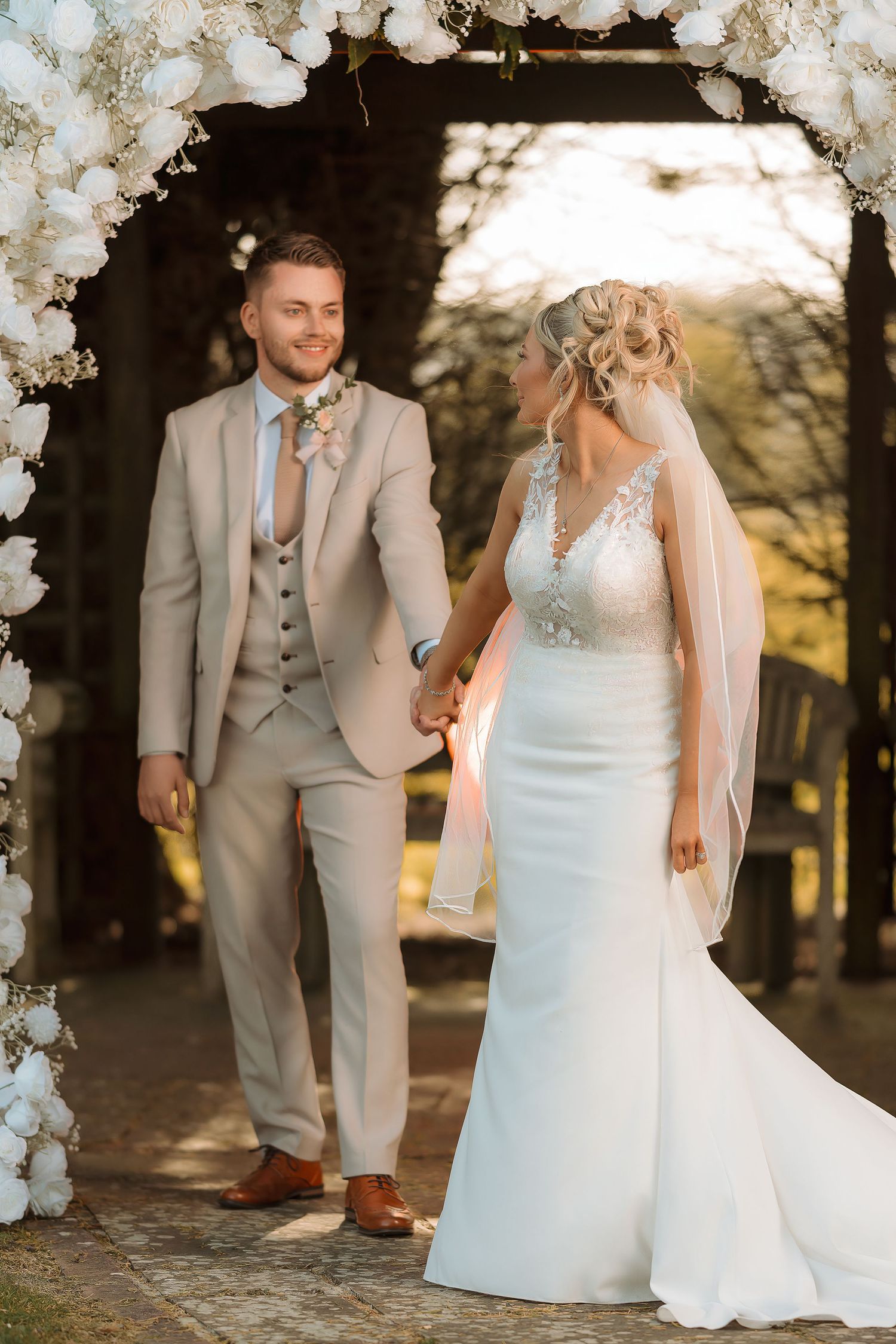A smiling bride and groom hold hands under a floral arch outdoors, dressed in a white gown and beige suit.