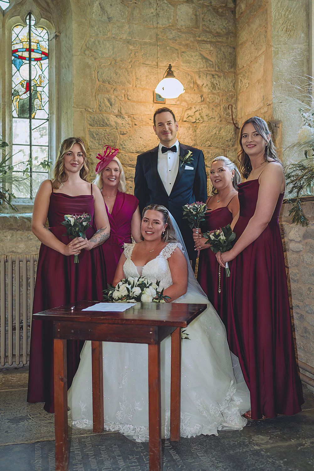 Bride seated at signing table with groom and three bridesmaids in burgundy gowns inside a stone church.
