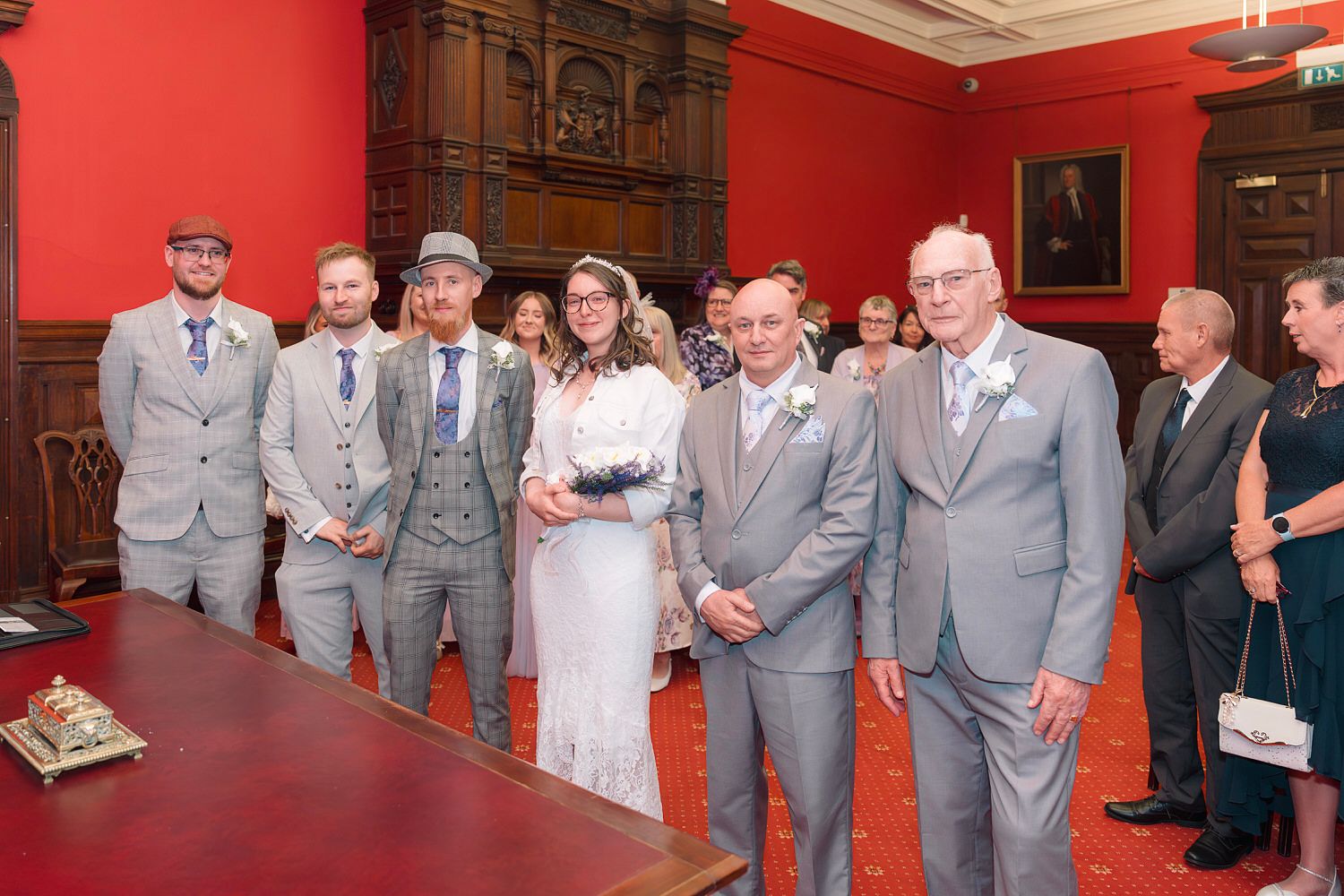 Wedding party posing in an elegant red-walled room, featuring bride in white dress with groomsmen in grey suits.