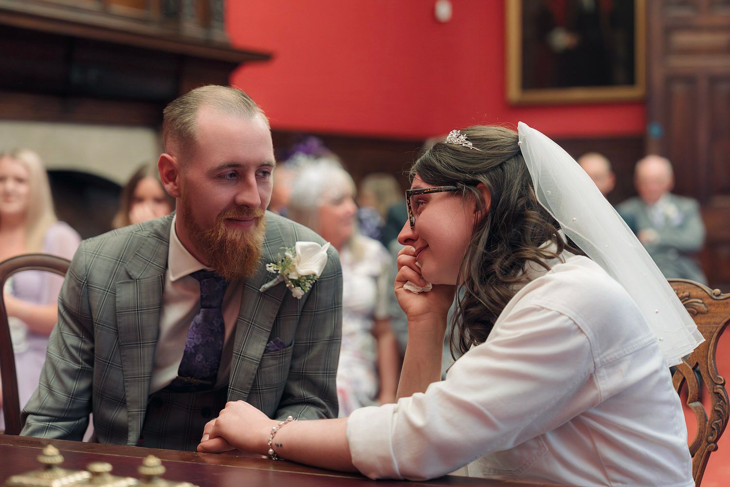 Bride and groom share an intimate moment at wedding ceremony table, bride in veil smiling at bearded groom in gray suit.
