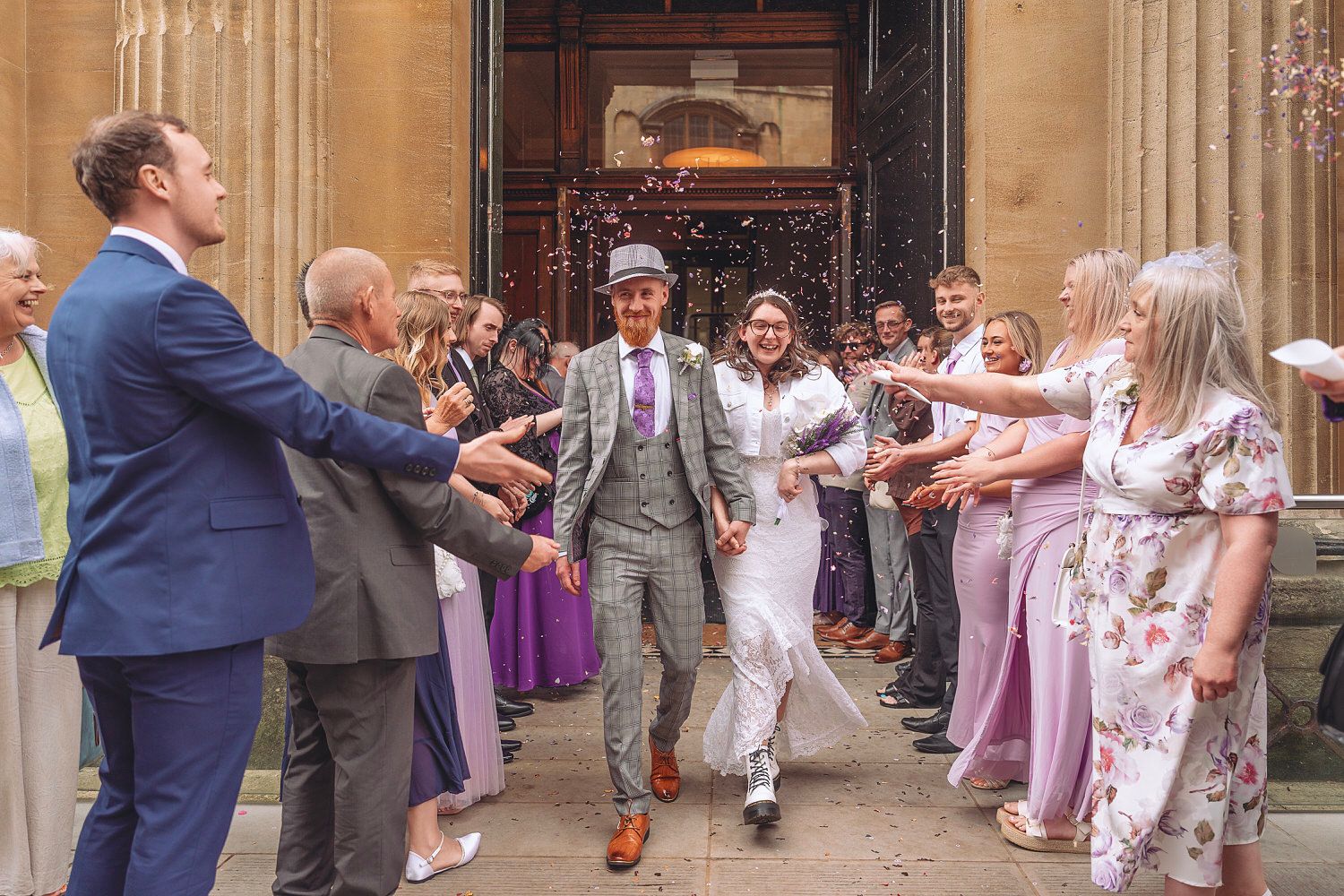 Joyful newlyweds exit church amid confetti shower, surrounded by cheering guests in purple and floral outfits.