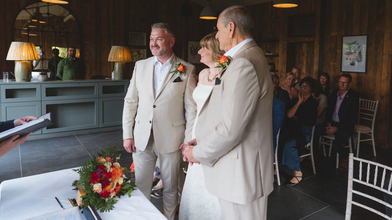 Couple exchanges wedding vows indoors with officiant and guests seated in rustic venue decorated with colorful flowers.