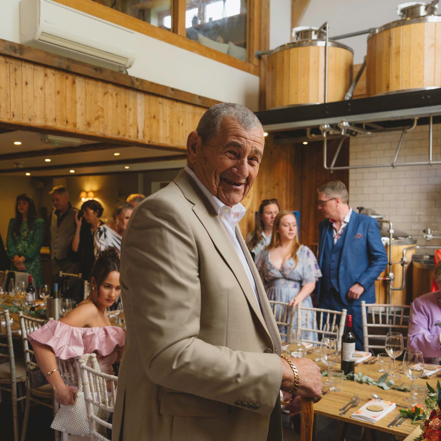 Elderly man in beige suit smiling at an indoor wedding reception in a rustic venue with guests seated at decorated tables.