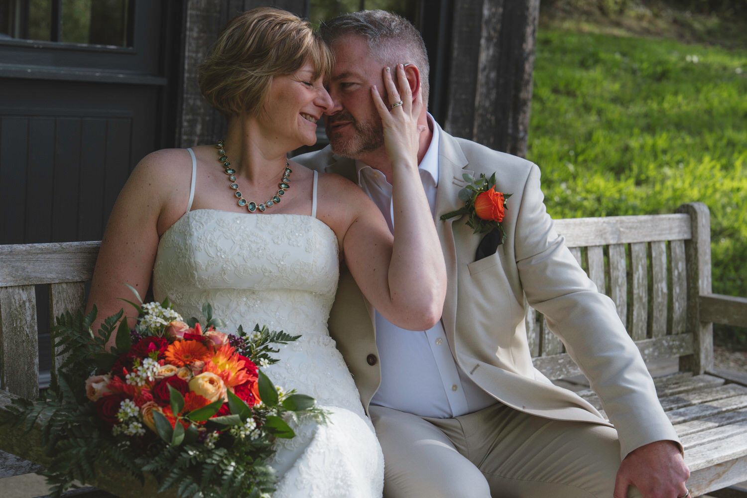 Bride and groom sitting on a wooden bench, sharing an intimate moment with a vibrant red and orange wedding bouquet.