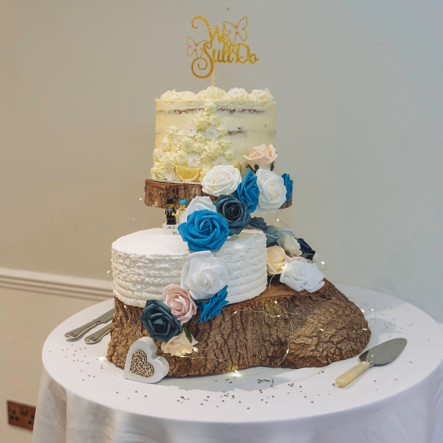 Three-tier wedding cake with teal and white roses on rustic wood base, topped with gold We Do topper.