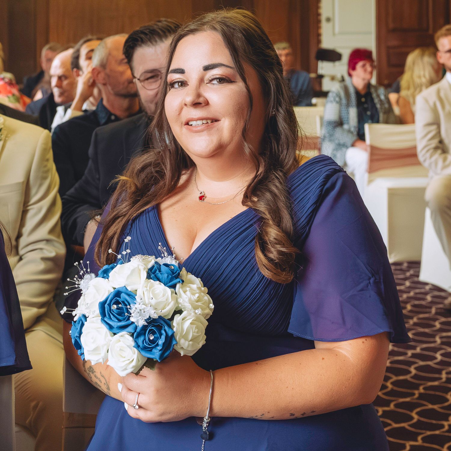 Woman in navy blue bridesmaid dress holding blue and white rose bouquet at a wedding ceremony.