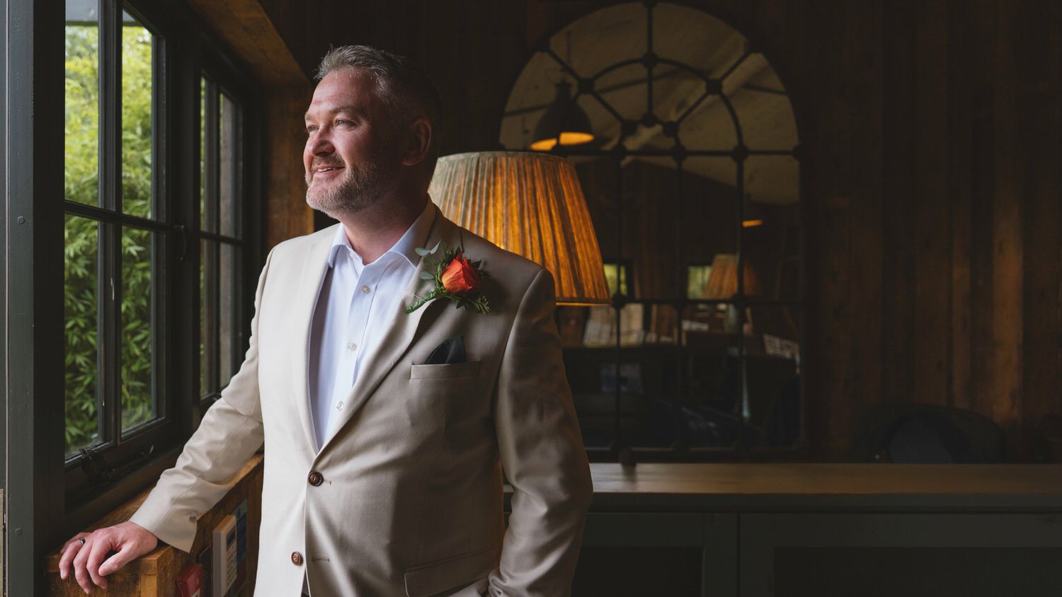 Groom in beige suit with floral boutonniere standing by window in elegant venue.