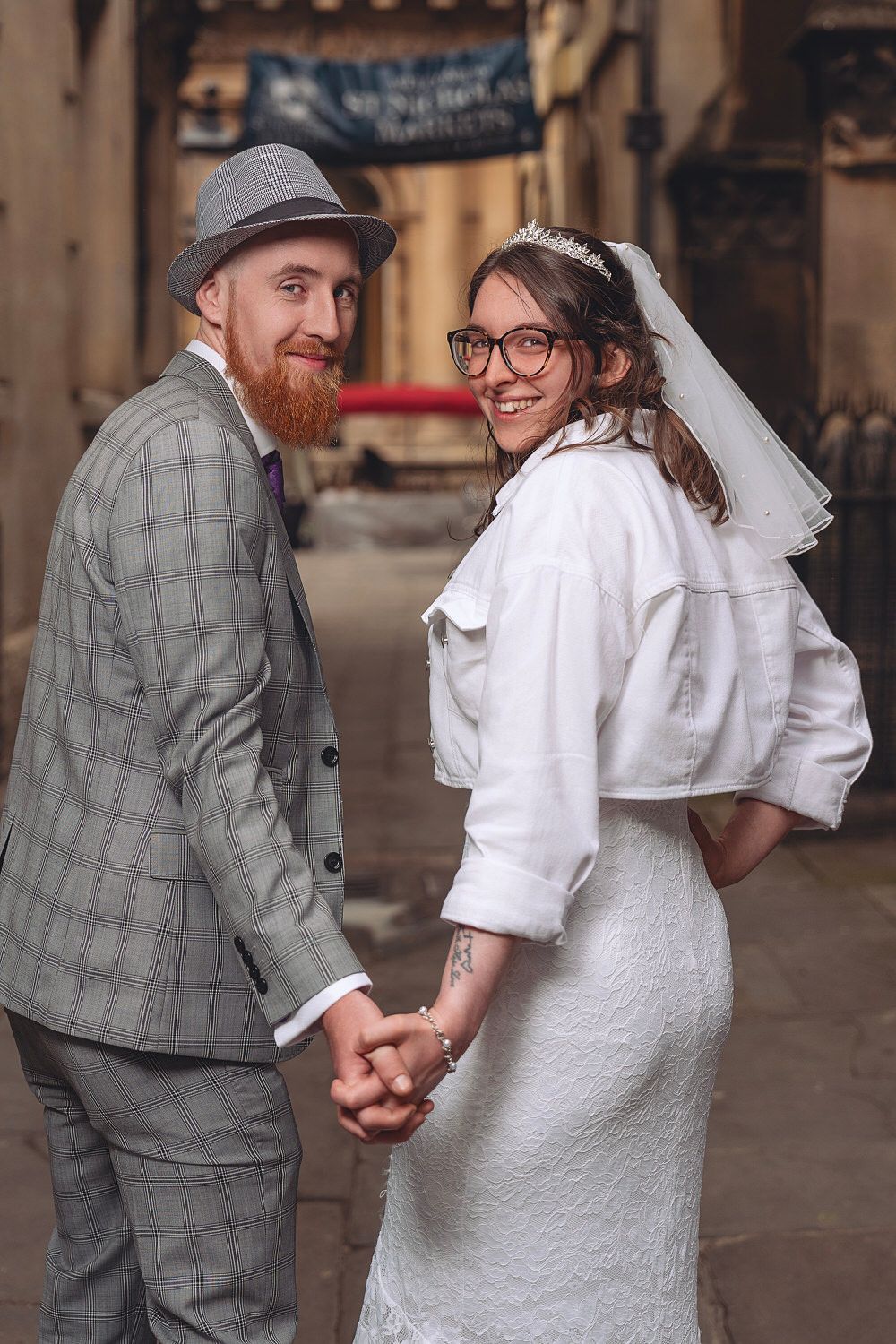 Smiling newlywed couple holding hands, bride in white lace dress and veil, groom in gray plaid suit and hat.