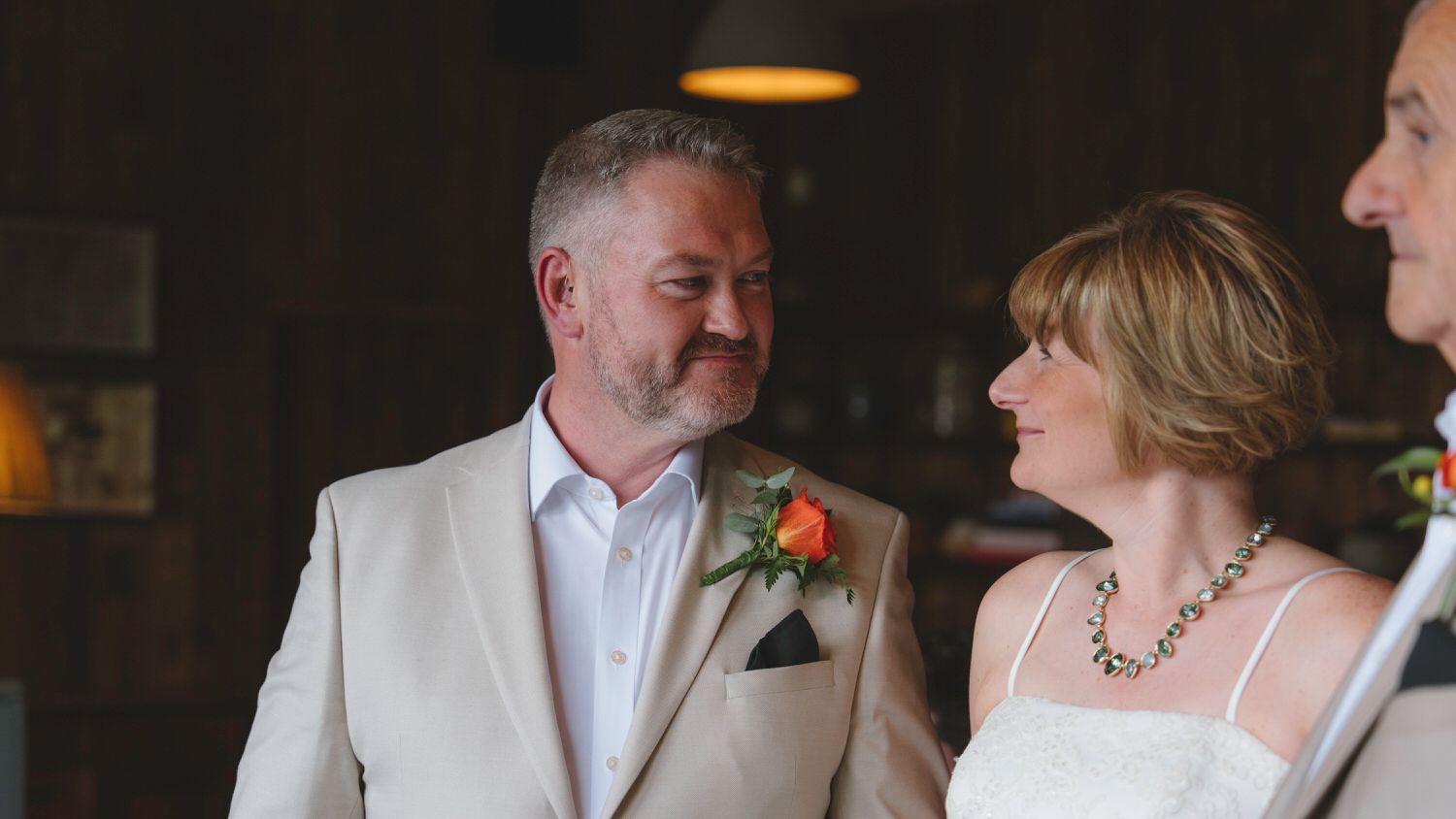 Groom in beige suit with orange boutonniere smiles at bride during intimate indoor wedding ceremony.